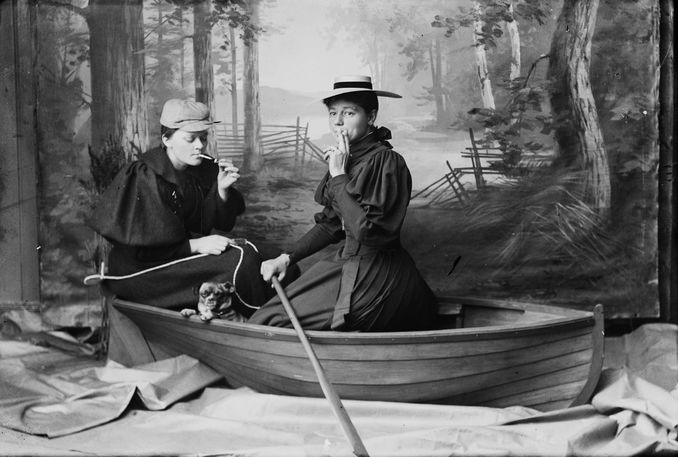 Studio portrait of two women and a small dog posed in a rowboat against a painted woodland backdrop with trees and fence.