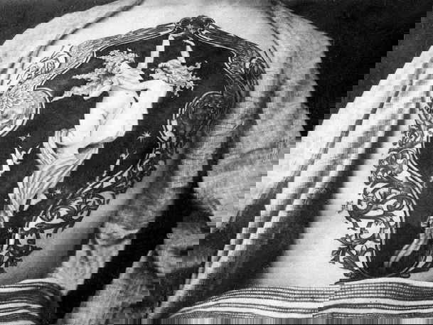 Exposed back shows large tattoo depicting two nude figures dancing among stars, surrounded by an ornate frame with flowing scrollwork designs.