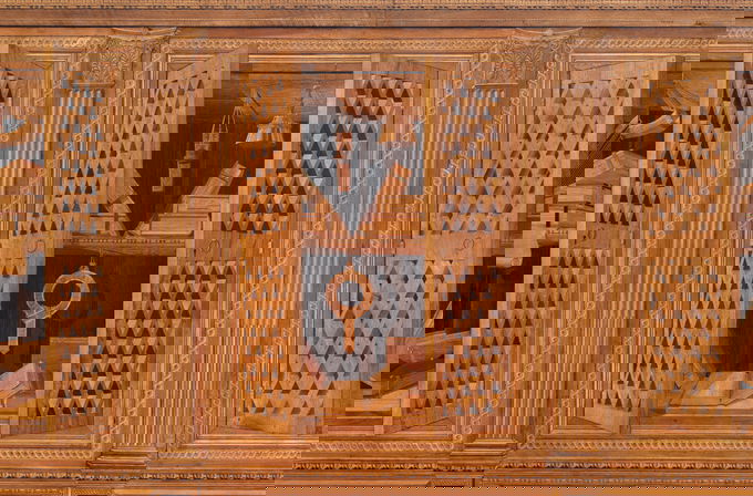 Intarsia depicting wooden cabinet with latticed doors revealing open books and hanging instruments inside, surrounded by ornately carved columns and decorative trim.