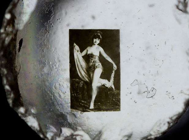 Microphotograph viewed through optical device showing a woman posing with draped fabric, surrounded by circular vignetting and visible dust particles.